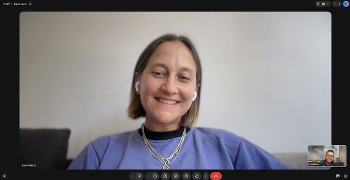 A woman with short brown hair, wearing a blue shirt and layered silver necklaces, smiles during a video call, with the name "Lenka Hierzi" visible in the bottom left and a small thumbnail of another participant, labeled "Gaurav Mohan," in the bottom right.