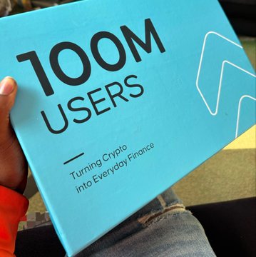 A person holding a teal box with the text '100M USERS' and 'Turning Crypto into Everyday Finance' printed on it.