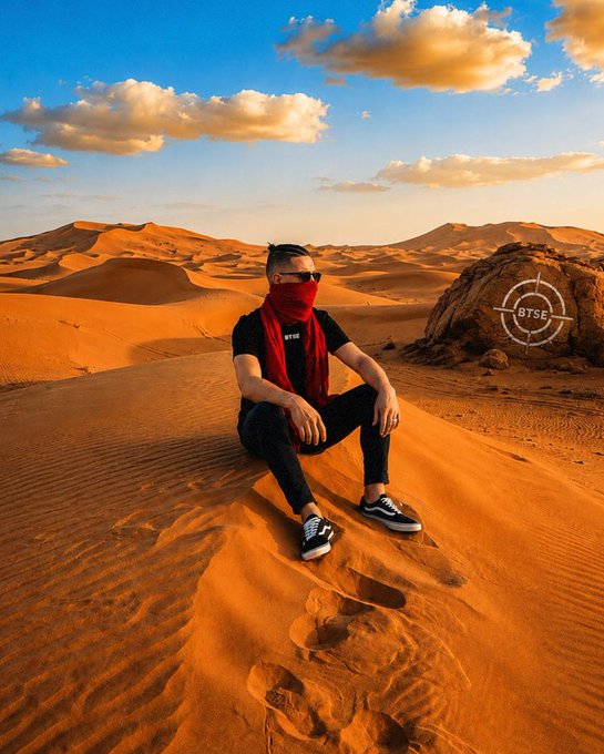 The image features a man sitting on a sand dune, wearing a red scarf around his neck and black pants. The background of the photo shows a desert landscape with mountains visible in the distance under a cloudy sky. There are also some clouds present in the foreground. The caption for this image reads "Focus on what is in the image and how it relates to #BTSE goes everywhere. Trade from anywhere trade.btse.com/join_btse".