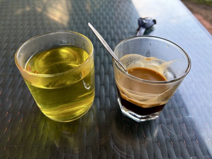 Two glasses, one with a yellow-green liquid and another containing espresso with a spoon, sit on a dark woven outdoor table with sunglasses in the background.