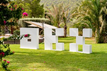 Large white sculptural letters spelling "FHE" are displayed on a grassy lawn with palm trees and flowers in the background.