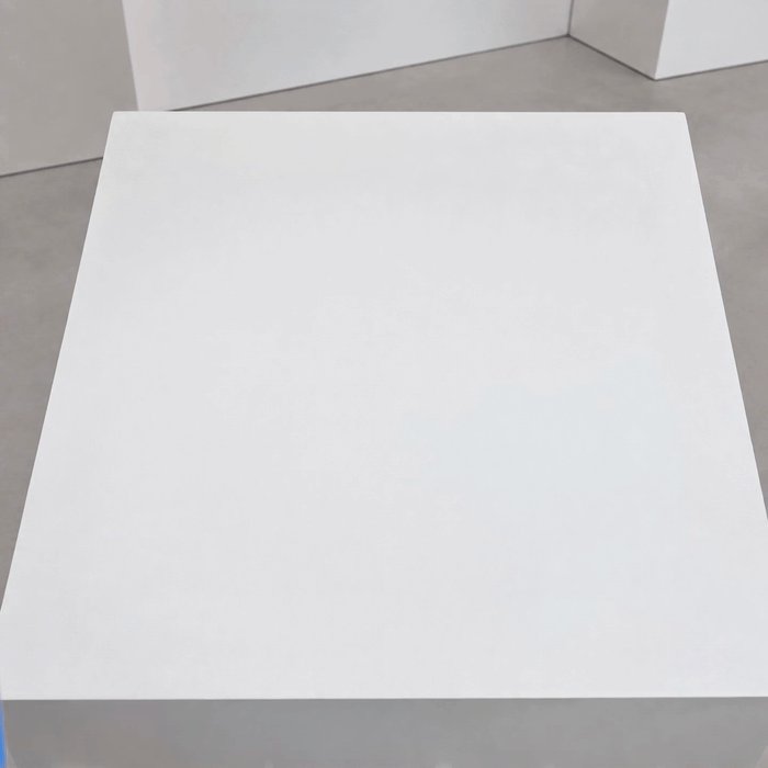 The image features a white square box placed on a gray floor. The box has a blank background or white surface that appears to be empty. In the background, there are two other boxes, one of which is also white but positioned at an angle. The main focus of the image is the white square box with no visible objects inside it.