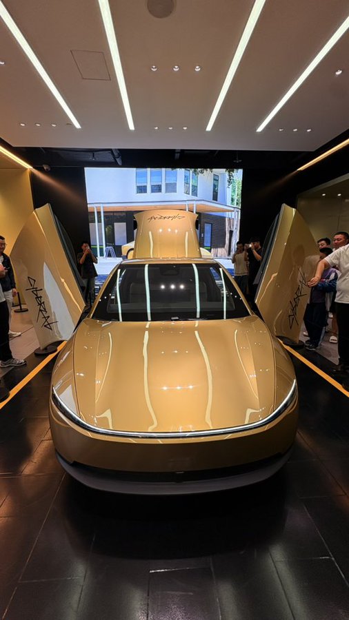 A gold-colored car displayed in a showroom with its hood open, viewed from the front under dramatic overhead lighting.
