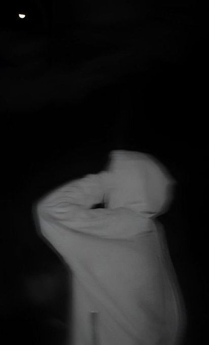 A blurry black-and-white nighttime photo shows a person in a light-colored hoodie with their hood pulled up, with only a small distant light visible in the dark background.