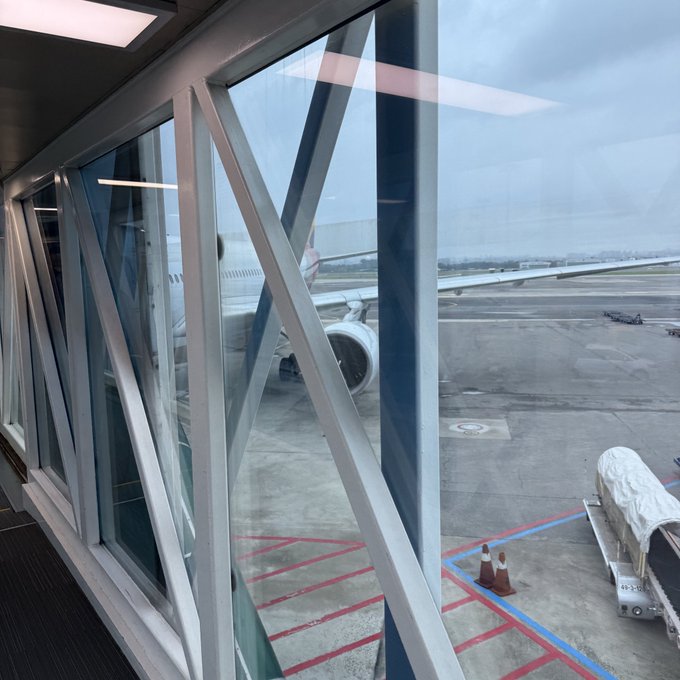 A view through a jet bridge window looking out onto an airplane parked on the tarmac under a cloudy sky.