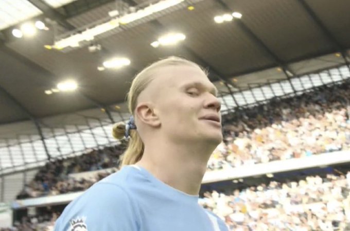 A close-up photograph of soccer player Erling Haaland wearing a light blue Manchester City jersey, smiling in a stadium filled with spectators.
