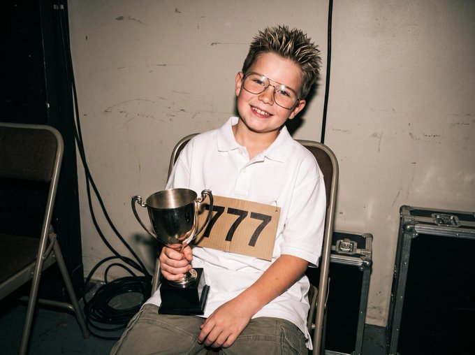 A young boy with spiky hair and glasses smiles while holding a silver trophy and a cardboard sign reading '777', seated in what appears to be a backstage area.