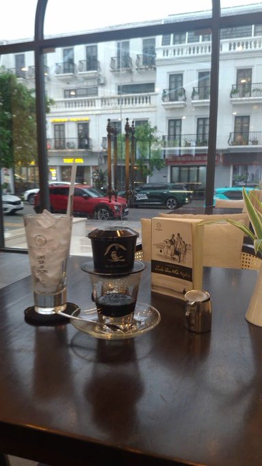 A Vietnamese coffee (cà phê sữa đá) served in a traditional phin filter over a glass of condensed milk and ice, accompanied by a glass of ice water, sits on a dark wooden café table by a window overlooking a Parisian street with classic Haussmann-style buildings and parked cars; the menu card reads "Cà phê Hoàng" and "Cà phê Rang Xay" with a rooster illustration, tying into the caption's call to start the day with coffee before getting to work ("Làm ly cafe rồi cày cuốc thôi nào").