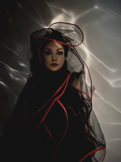 An artistic portrait of a woman wearing a black hat and flowing black veil with red ribbon accents, set against a dramatic white and gray backdrop.