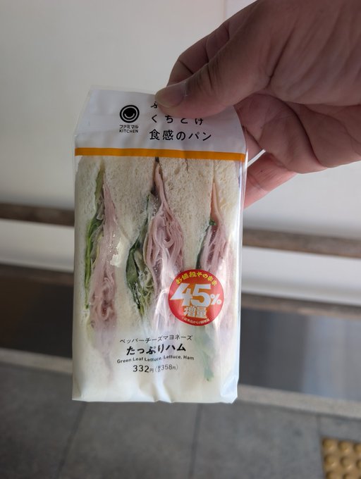 A hand holding a packaged Japanese sandwich from 7-Eleven ("つくる、食感のパン") containing Green Leaf Lettuce, Lettuce, and Ham, advertised as 45% larger.