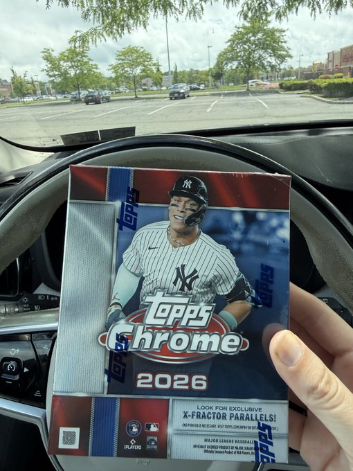 A hand holds a sealed 'Topps Chrome 2026' baseball card hobby box in front of a car steering wheel, featuring Aaron Judge on the cover and a 'LOOK FOR EXCLUSIVE X-FRACTOR PARALLELS!' sticker.