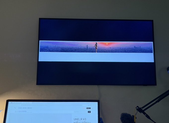A photograph of a workspace showing a wall-mounted television displaying a cinematic video of a person overlooking a city at sunset, with a laptop below showing a webpage with Thai text "UND P #3."