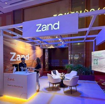 A Ripple event booth prominently displays the name “Zand,” with a reception desk, lounge chairs, and branded signage. The image aligns with the post’s description of Zand as an AI-powered bank for the digital economy, in a Ripple-focused “Looking for ‘Z’?” setting.