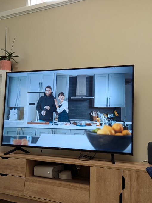 A living room scene shows a flat-screen TV playing a cooking show with two people in a kitchen, sitting on a wooden media console with a router beneath it.