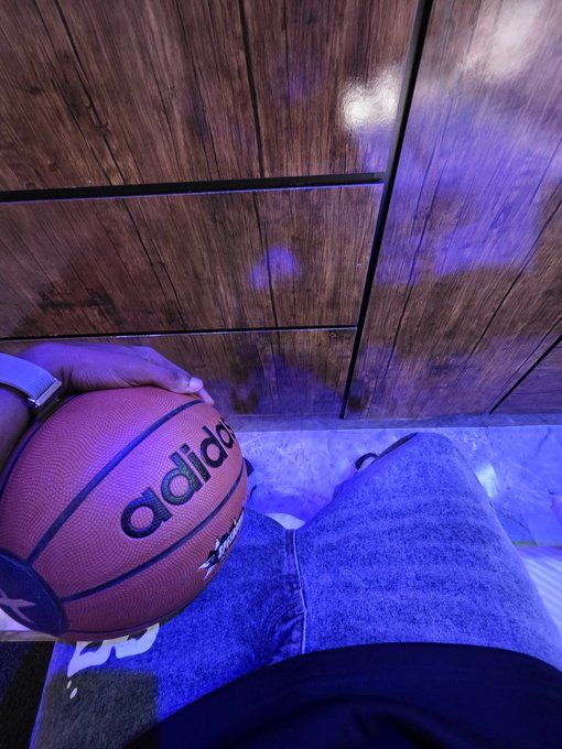 A close-up shot of a hand holding an Adidas basketball on a wooden floor illuminated by purple lighting.