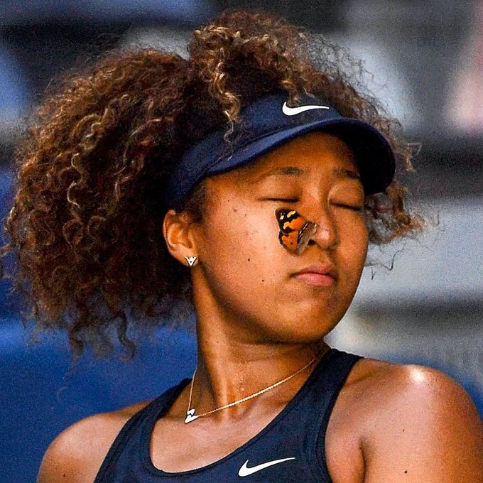 Tennis player Naomi Osaka with curly hair wears a navy Nike visor and athletic top, with butterfly tape on her nose and eyes closed.