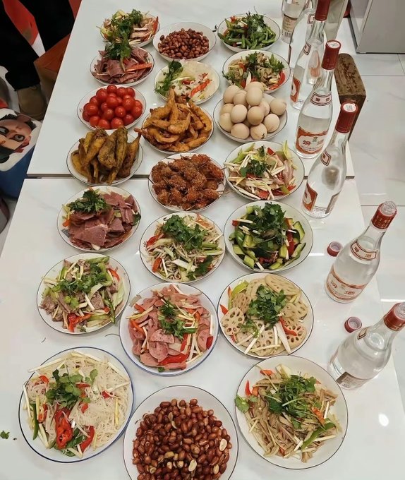 A table covered with numerous plates of Chinese dishes, including peanuts, tomatoes, eggs, shrimp, and various stir-fries with cilantro, accompanied by bottles of liquor.