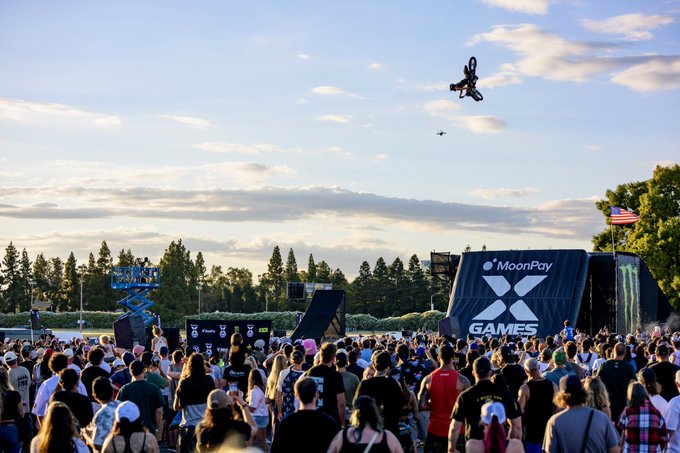 A motocross rider performs an aerial stunt high above a large crowd at the X Games, with visible signage reading "MoonPay" and "X GAMES" on the ramps below. The caption "MOON THAT" plays on the MoonPay branding while referencing the rider's soaring, moon-bound trajectory through the sky.