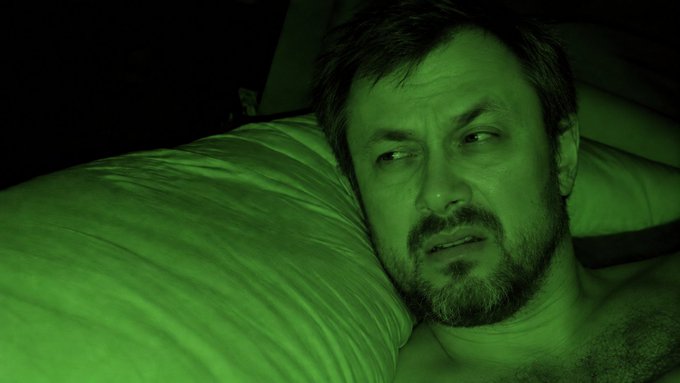 A close-up, green-tinted photograph of a bearded man lying on a pillow, gazing off to the side with a pensive expression in dim lighting.