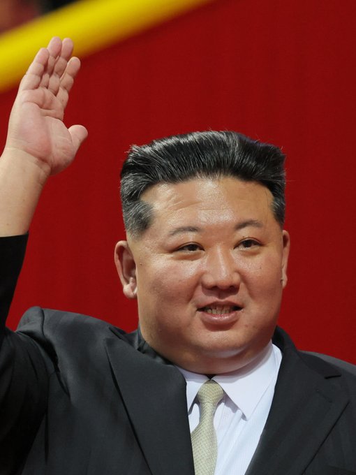 Kim Jong Un waves his hand while wearing a suit and tie against a red background.