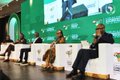A panel discussion on a stage with a green backdrop showing two seated speakers, appearing to be at a conference or summit event.
