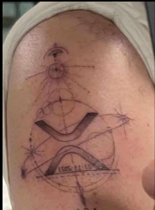 A geometric tattoo on a person's shoulder featuring a stylized "X" with celestial elements like stars and a sun, with the text "LUCAS €1.07" written at the bottom, humorously depicting a price tag for the artwork. The "$1.07" caption pokes fun at the bargain-basement pricing of this elaborate celestial design.