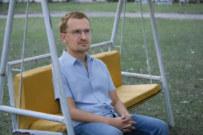 The image shows a man sitting on a swing, wearing glasses and a blue shirt. The swing has yellow padding for comfort. In the background, there are chairs and benches scattered around the area, suggesting that this might be an outdoor seating area or a park setting. A caption in the image reads "i feel sorry for the people who bought the bottom of the bottom of $gitlawb and many other top coins hope @jessepollak will pump everything soon".