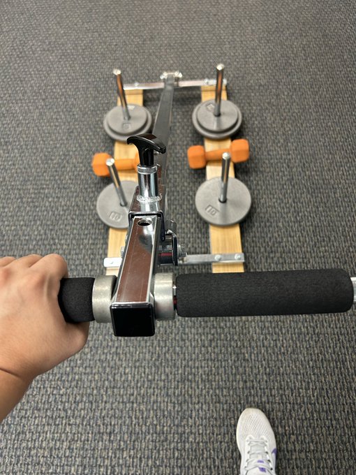 A first-person view of a hand gripping the handles of a metallic glute/hamstring developer machine with weight plates on a carpeted floor.