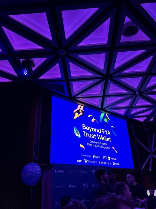 A large screen displays 'Beyond 9YA Trust Wallet TOKEN2049 | Singapore October 6 | 6:10 PM' under a dramatic purple-lit geometric ceiling at an event.