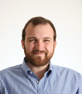 The image shows a smiling man with a reddish beard wearing a blue and white checkered button-up shirt, likely Charles Hoskinson, the Cardano founder referenced in the post warning about the compromised IOG YouTube channel.