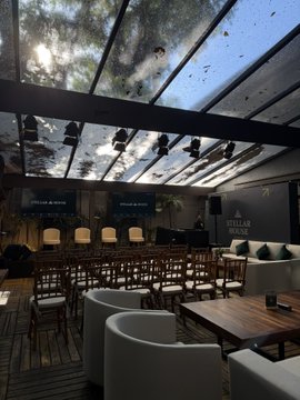 An elegant indoor-outdoor event space features rows of wooden chairs arranged beneath a glass canopy adorned with hanging greenery, with "STELLAR HOUSE" signage visible in the background. The sophisticated venue setup accommodates the Stellar House SP event hosted by StellarOrg and rivool_finance, providing an intimate atmosphere for attendees.