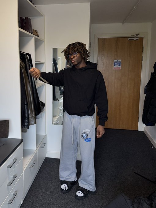 A young man with glasses and dreadlocks poses in a bedroom closet, wearing a black hoodie, gray BMW Motorsport sweatpants, and slides. The caption's casual tone and reference to "zaddrnet" suggests the photo is part of a promotional post seeking whitelist access, likely for an NFT project.