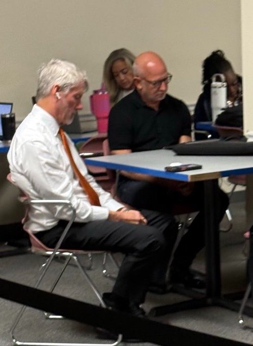 Two men sit side by side at a conference table, both looking down intently, seemingly engrossed in or waiting for something off-screen—capturing the patient, focused anticipation of "waiting for the bull run."