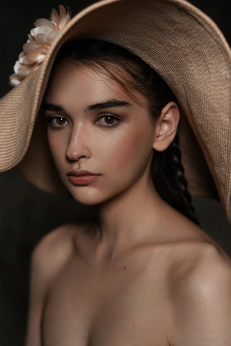 A portrait of a young woman with dark hair wearing a wide-brimmed beige sun hat adorned with a beige flower.