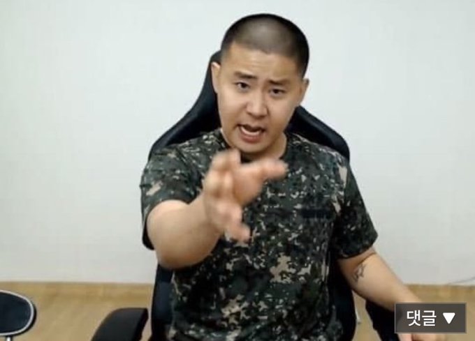 A man in a camouflage t-shirt sits in a black gaming chair, pointing an accusatory finger directly at the camera with an intense, animated expression. The image visually reinforces the post's urgent, confrontational tone about Bitcoin's predicted rise to 150k, capturing the speaker mid-rant as if calling out the viewer for not investing now. A Korean comment button "댓글" (Comments) appears in the bottom right corner.