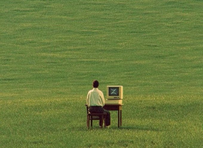 An artistic photo showing a person in a white shirt sitting at a desk with a vintage computer monitor in the middle of a vast green grass field.