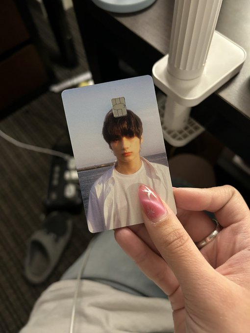 A hand with long pink decorated nails holds a photo card of a young man standing by the sea, clipped at the top with a small metal clip.