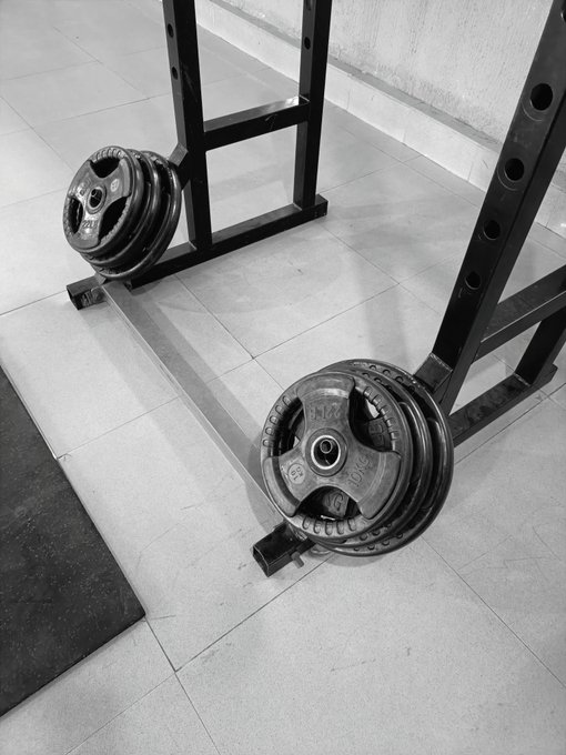 Black-and-white gym scene showing two barbells loaded with weight plates on squat racks beside a floor mat. The accompanying post claims **$INJ** doubled this week and says the clip is being shared in that context.