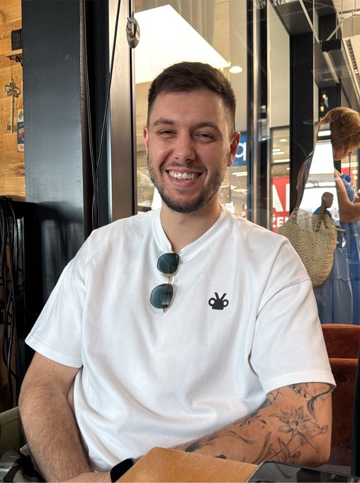 A smiling young man with a beard, wearing a white t-shirt with sunglasses hanging from the collar, sits at a table in a cafe with a tattoo visible on his forearm.
