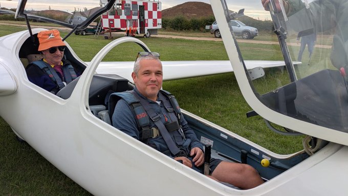Two men sit in a white glider on a grass airfield, smiling at the camera, with another glider and a red and white vehicle visible in the background.