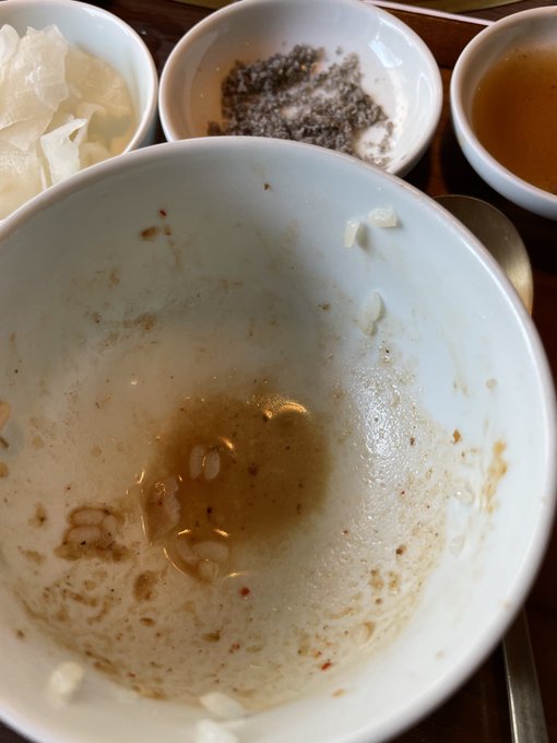 A nearly empty white bowl with food residue and a small puddle of liquid sits next to small side dishes containing remnants of white pickled vegetables, dark seasoning, and tea.