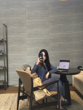A woman in a gray knit outfit sits in a stylish chair, taking a mirror selfie with her phone while holding a Chanel quilted handbag, with a laptop and a small side table beside her. The image reflects the "富婆姐妹" (rich lady sisters) theme by showcasing luxury and sophistication through the designer bag and elegant setting.
