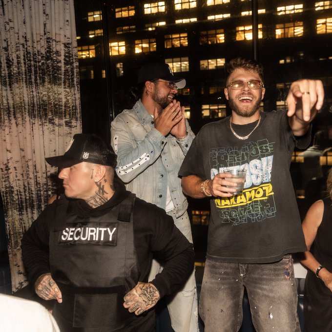 Two men laugh at a nighttime event with city lights in the background; one wears a denim jacket and cap while the other wears a black graphic t-shirt and glasses, standing near a man in a 'SECURITY' vest.