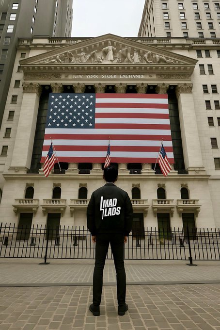 A person wearing a black jacket with 'MAD LADS' on the back, standing in front of the New York Stock Exchange building draped with a large American flag.