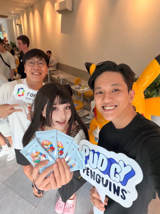 Three people at what appears to be a Pudgy Penguins event, with one holding trading cards and another holding a 'Pudgy Penguins' branded sign.