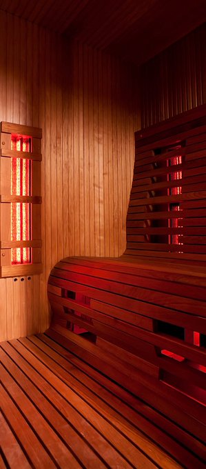 The interior of a wooden sauna is bathed in warm, reddish light, featuring cedar paneled walls, a wooden bench, and glowing infrared heater panels.