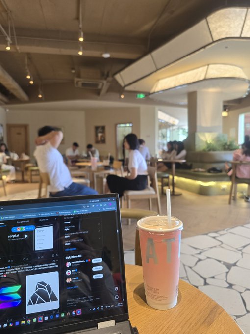An indoor cafe or co-working space scene shows a laptop screen and a pink drink cup on a wooden table in the foreground, with patrons seated at tables and a person stretching in the background.