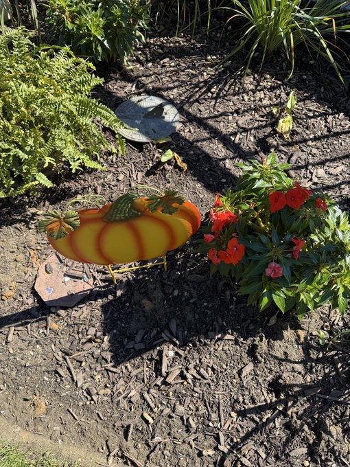 A garden scene shows decorative yellow and red striped chicken ornaments and a flowering red plant set in mulched soil among greenery.