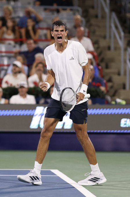 A tennis player in a white Adidas shirt and dark shorts celebrates with clenched fists on a blue tennis court.