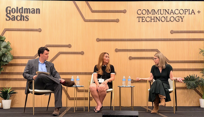 Three people seated on a stage during a panel discussion with 'Goldman Sachs' and 'COMMUNACOPIA + TECHNOLOGY' logos displayed on the wooden backdrop.